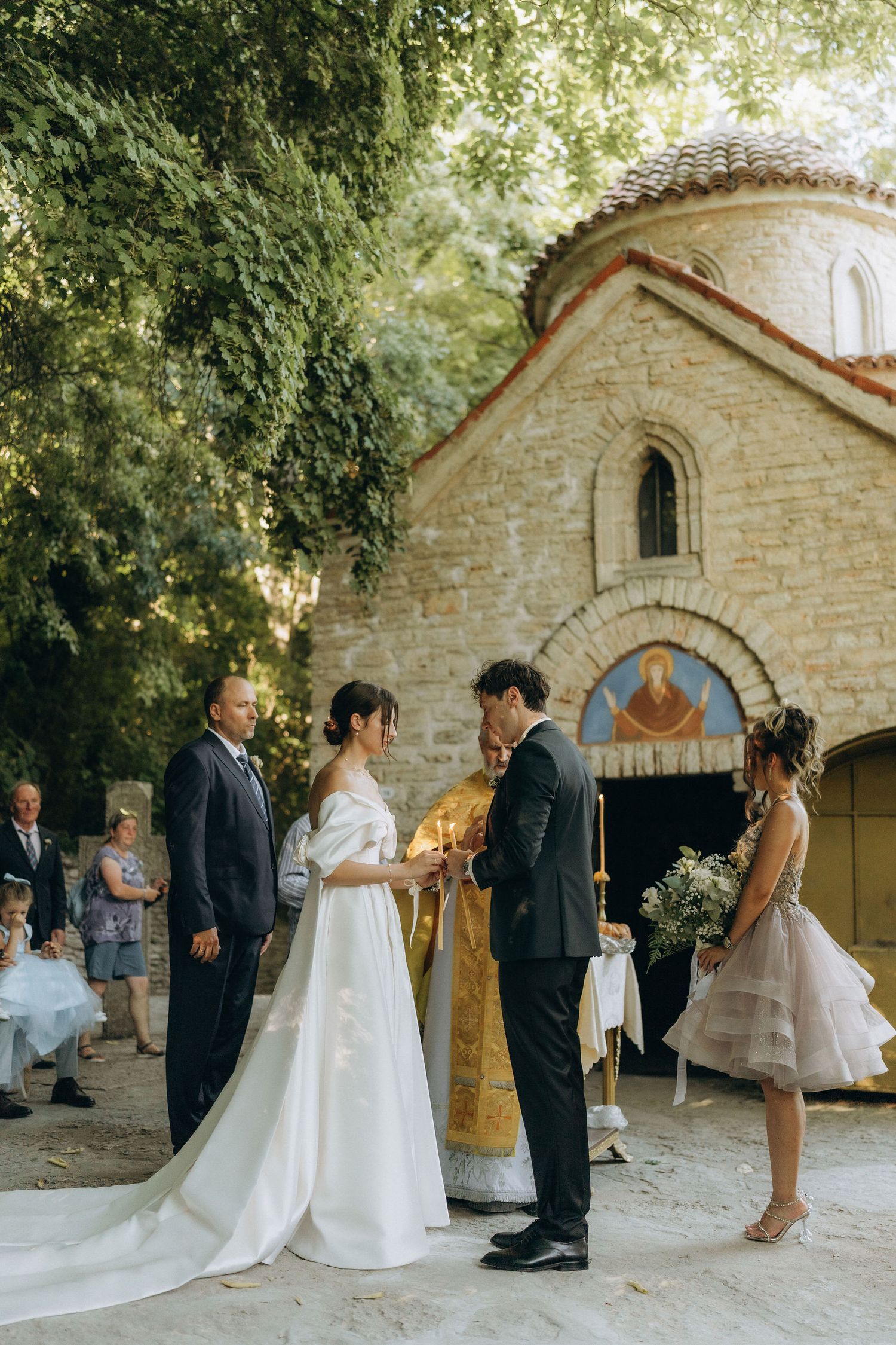 Wedding ceremony taking place outside a rustic stone chapel with guests gathered under green trees.