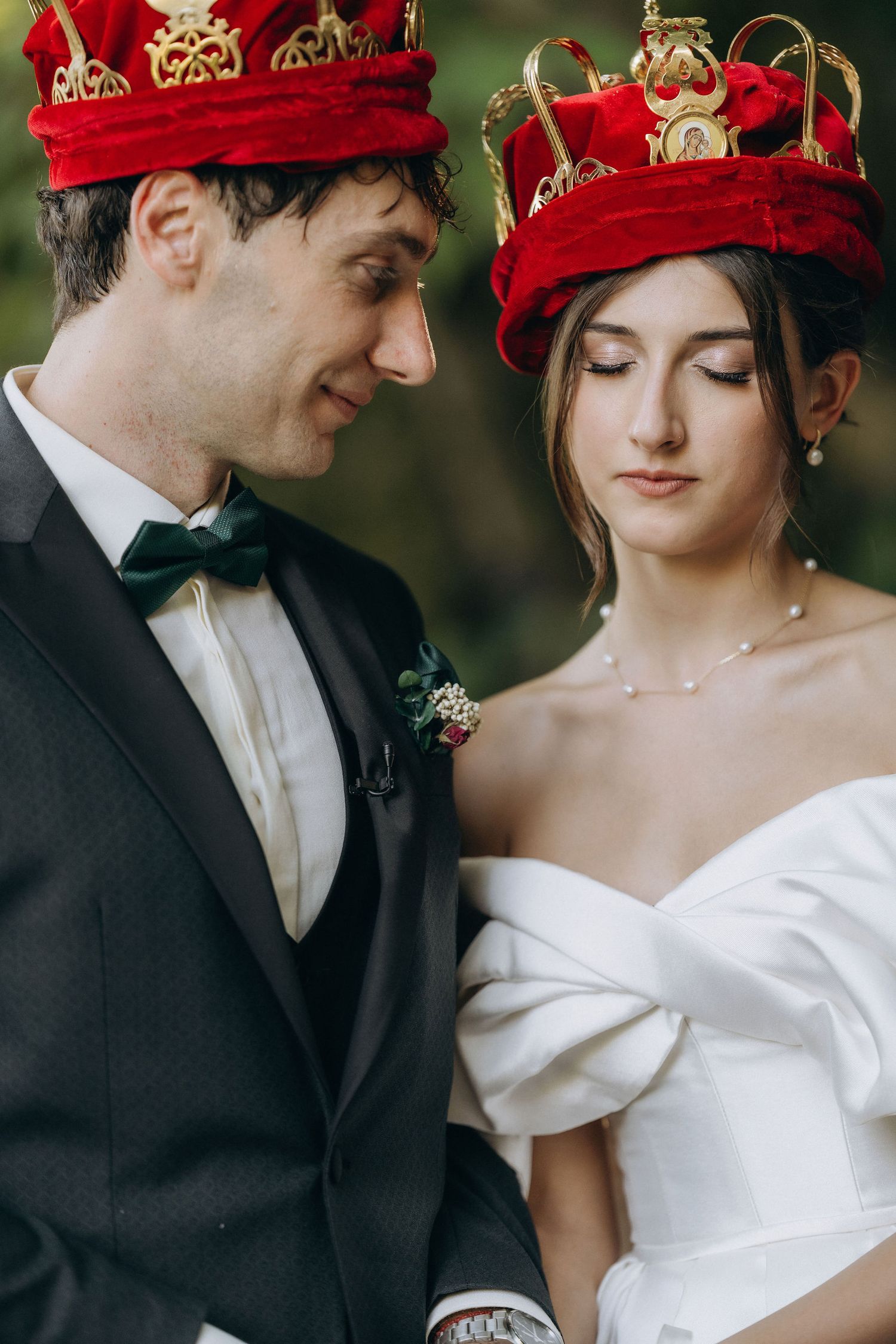 Couple in wedding attire wearing matching red velvet crowns with gold details.