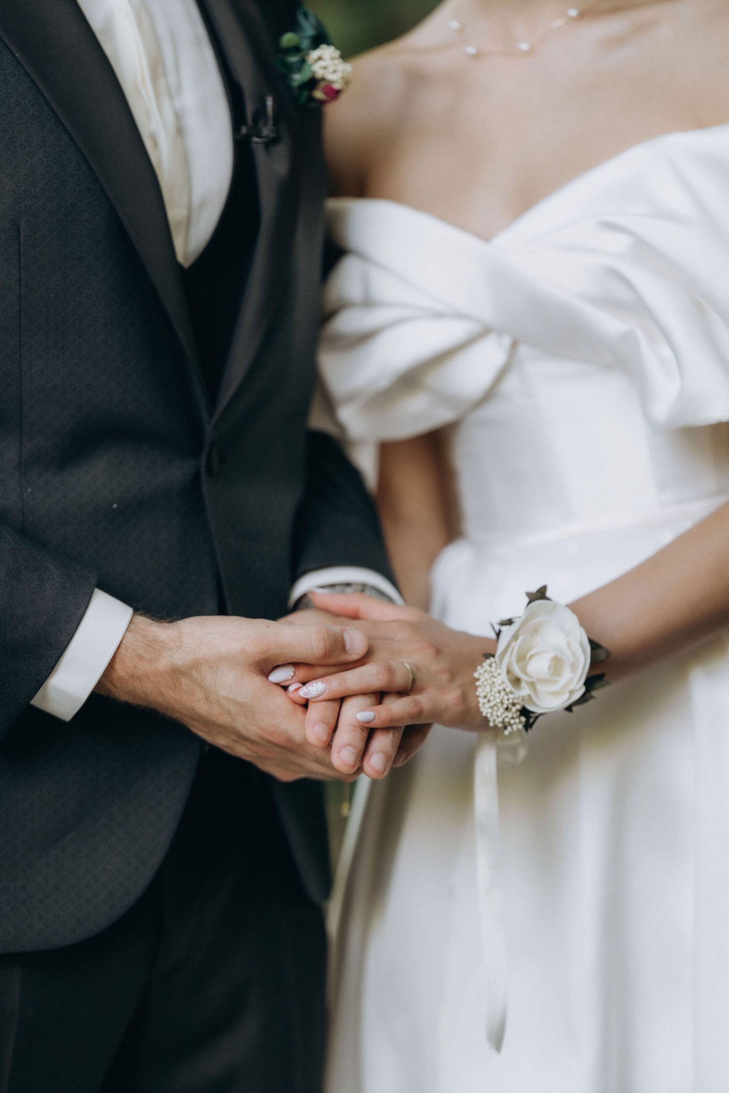 Close-up of bride and groom's hands clasped together, with white wedding dress and black suit details visible.