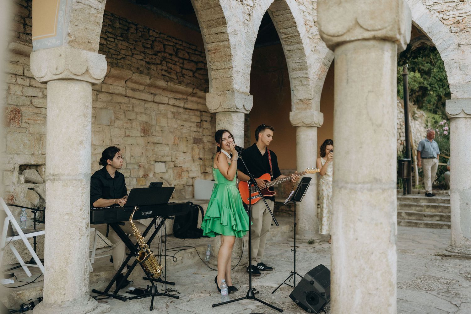 Stone colonnade and arches frame an outdoor wedding gathering.