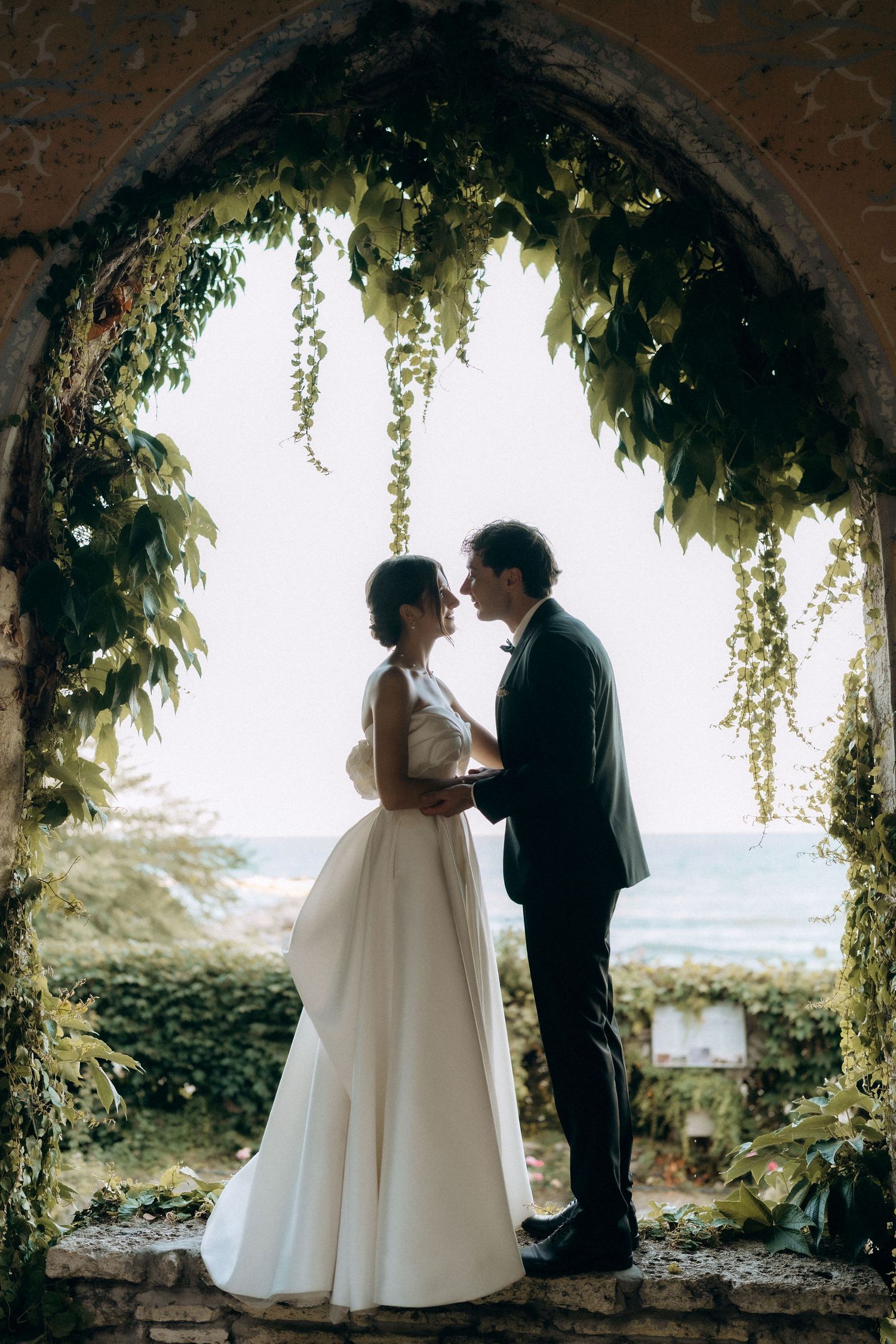 Wedding couple silhouetted against bright sky in an ivy-covered stone colonnade.