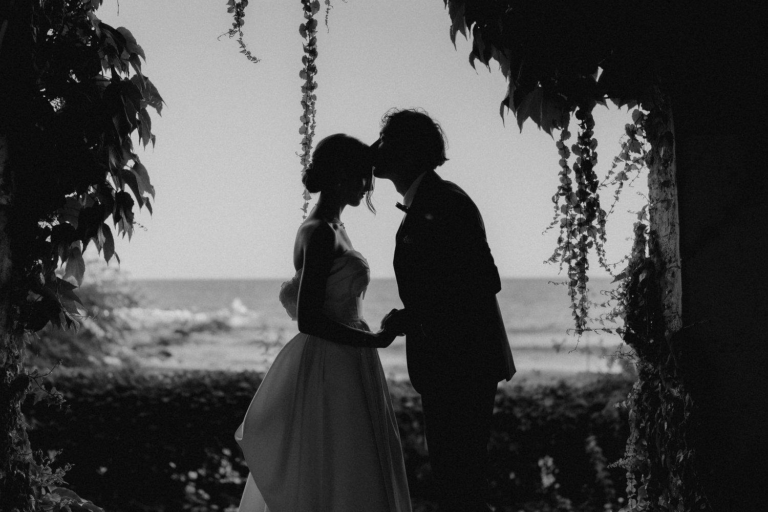 Black and white silhouette of newlyweds embracing under hanging vines.