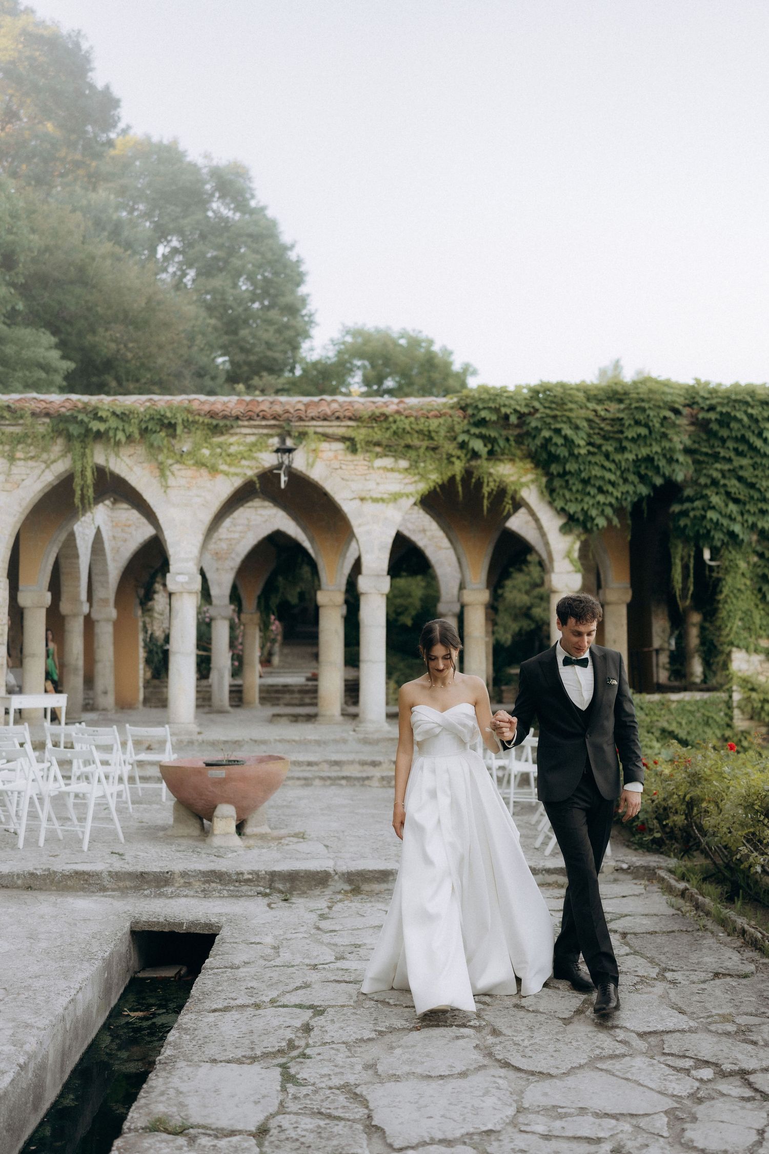 Wedding couple walking through a stone colonnade with white chairs set up for ceremony.