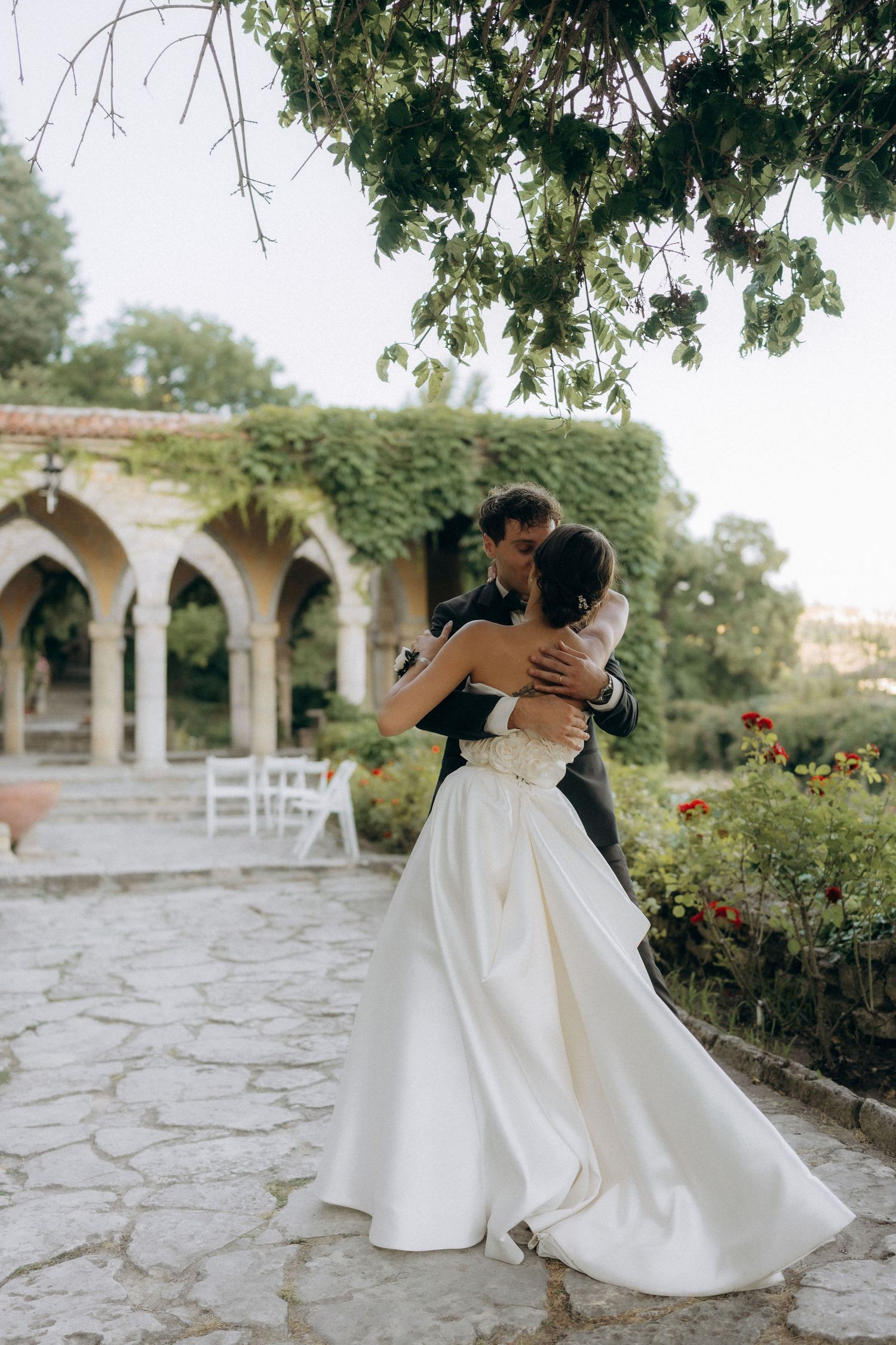 Newlyweds share an intimate dance on stone patio surrounded by arched columns and greenery.