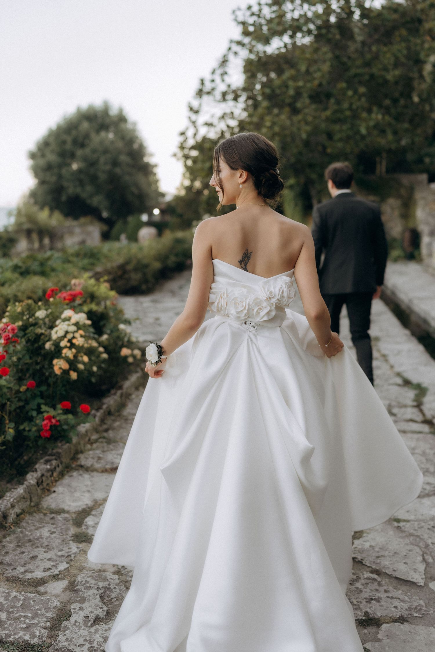Elegant bride in white strapless gown walks along stone path with red flowers and greenery in garden setting.