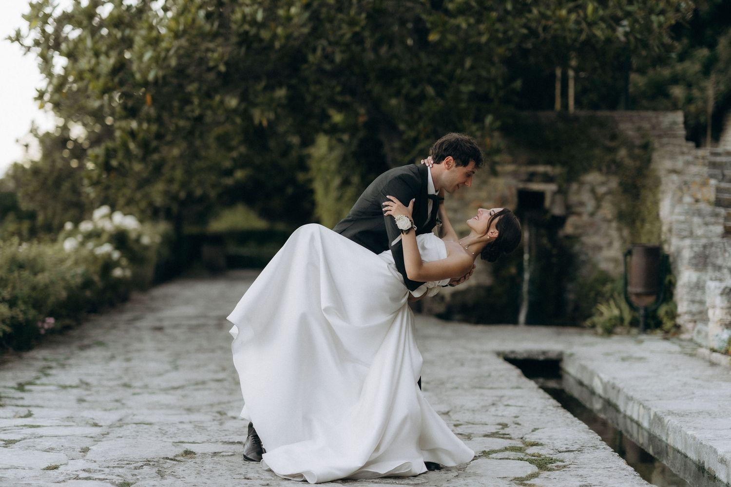 Wedding couple embracing on stone pathway surrounded by garden architecture.