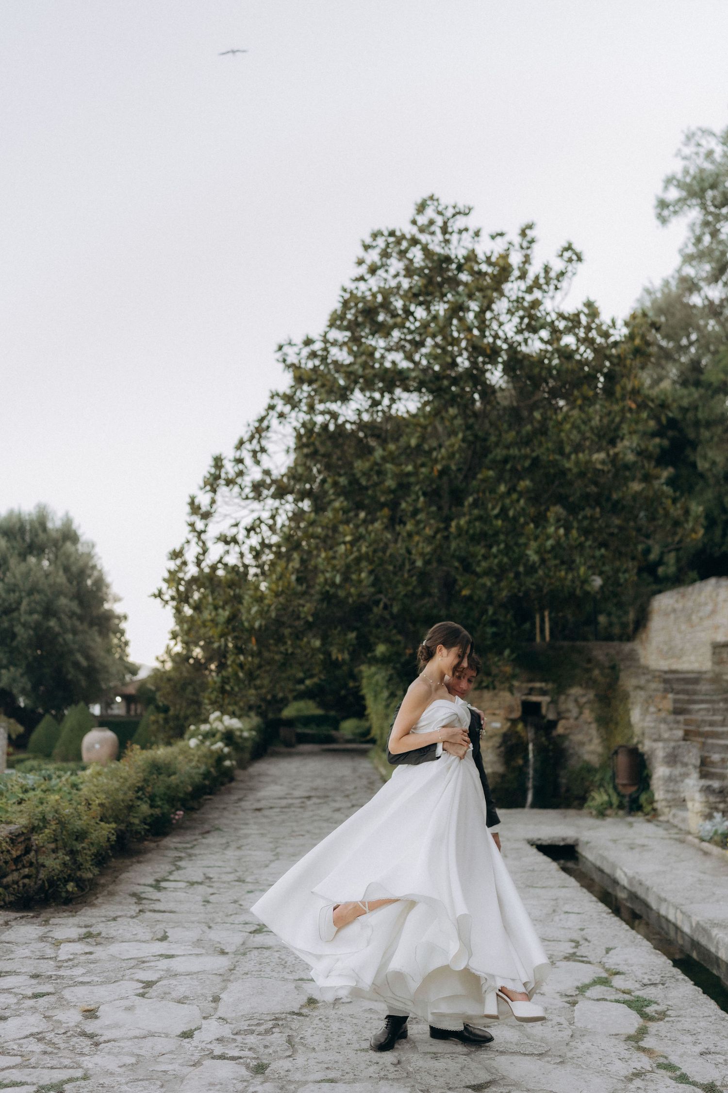 Bride in flowing white dress twirls on stone garden path near flowering bushes.