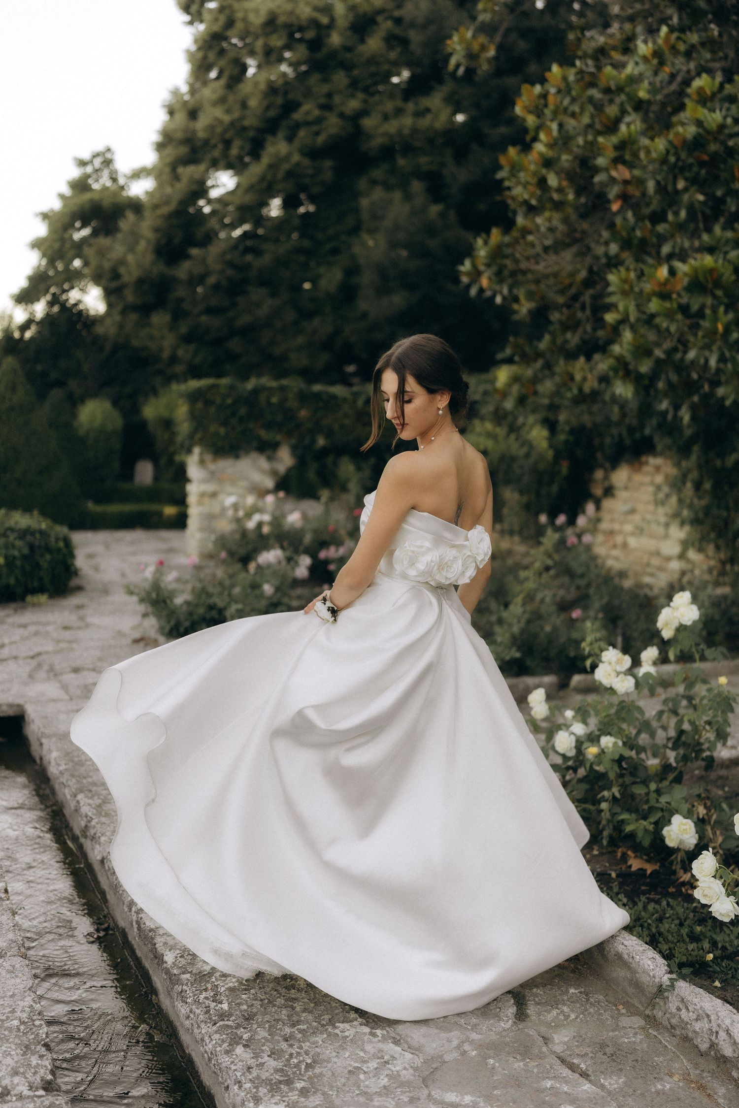 Bride in strapless ballgown poses gracefully on stone wall surrounded by white roses and greenery.