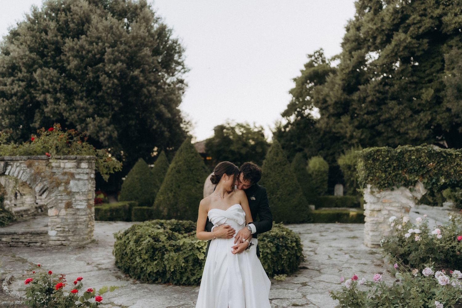 Newlyweds share tender moment in garden setting with stone walls and colorful flowers.
