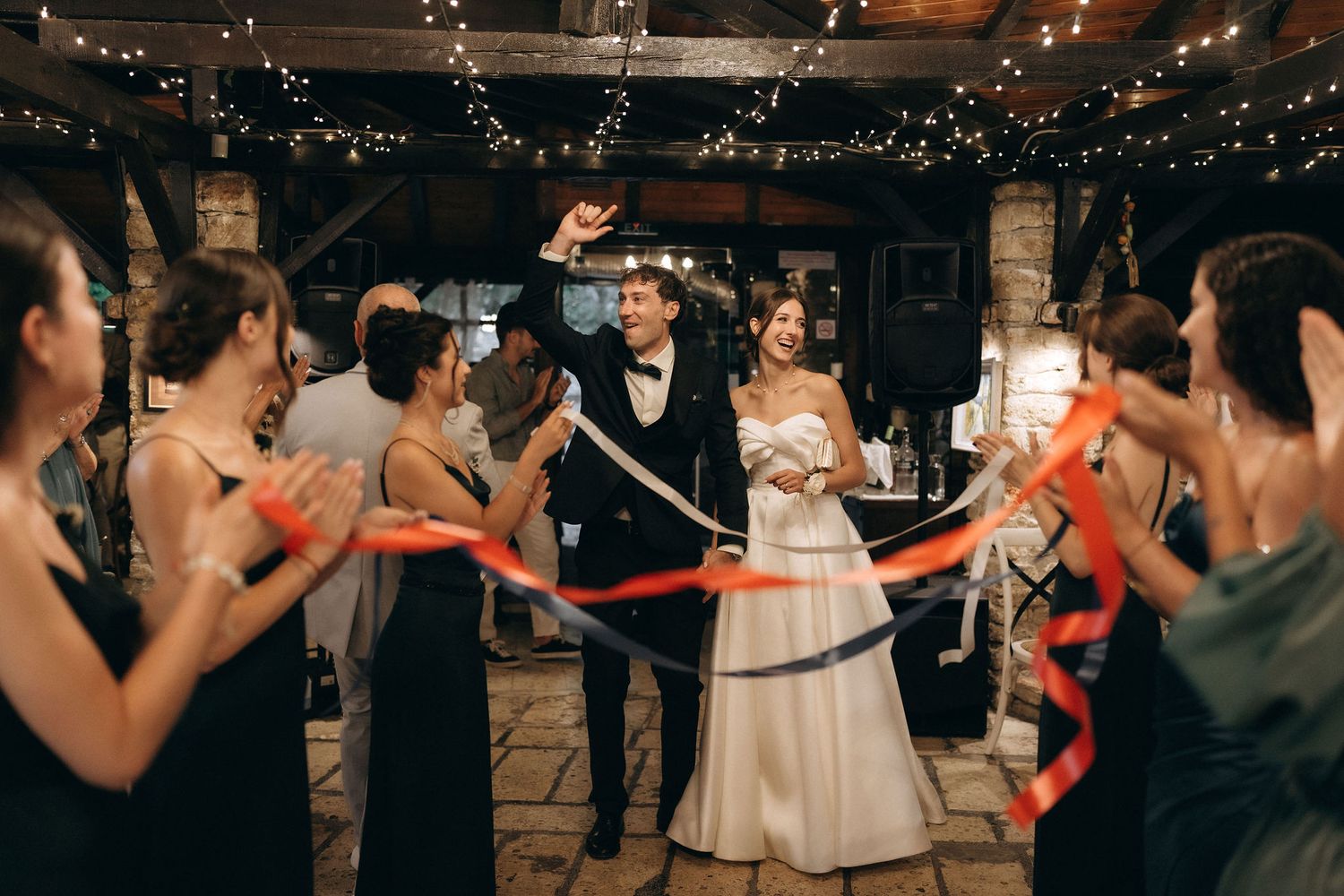 Wedding party members hold red ribbons during a reception celebration under twinkling lights.