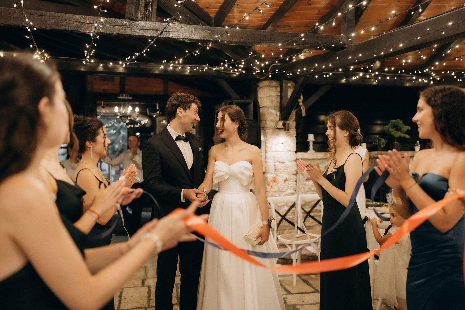 Wedding celebration in a rustic venue with string lights and stone columns as guests hold ribbons.