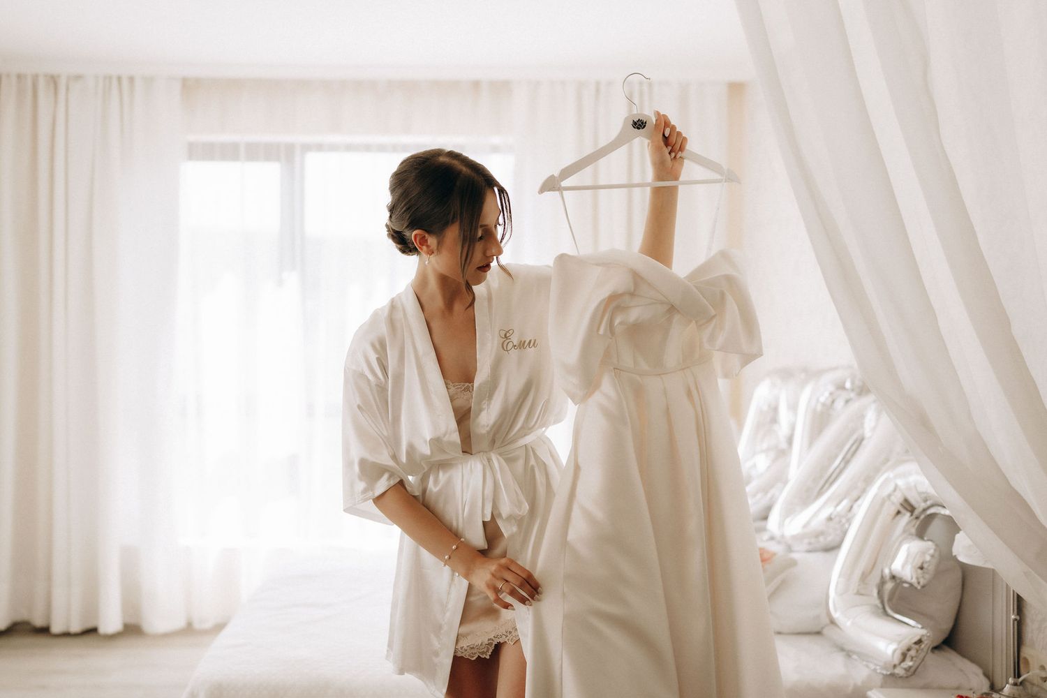 A bride getting ready in a bright white room with flowing curtains and natural light.