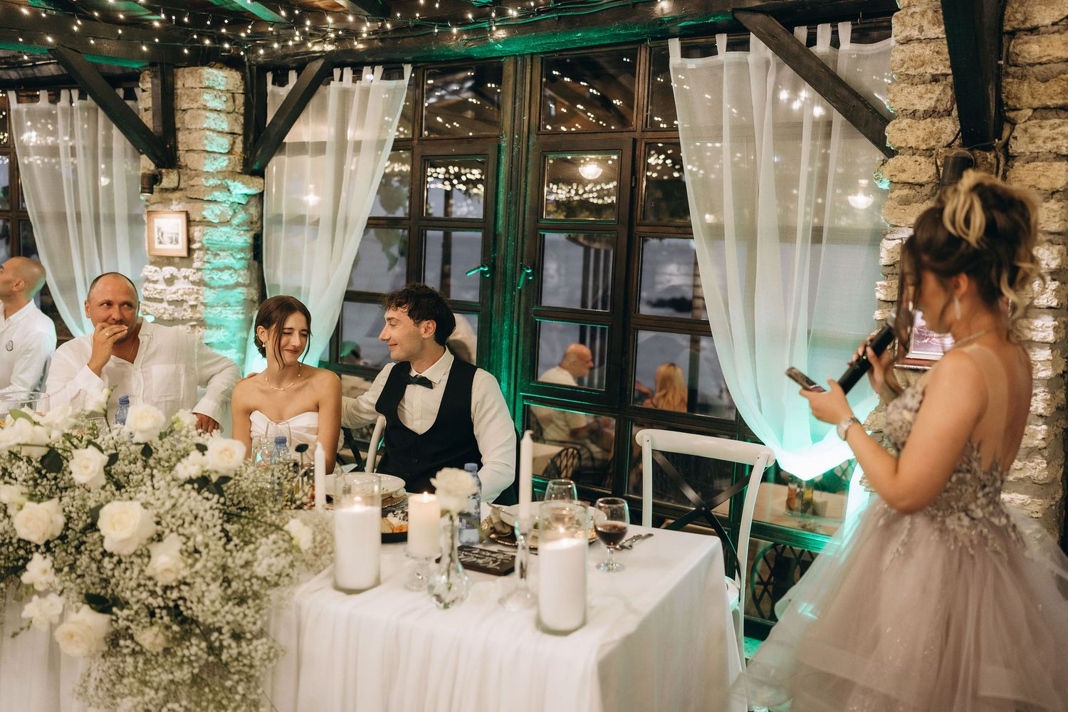 Wedding reception table setup with white flowers and decor in a rustic venue with string lights.