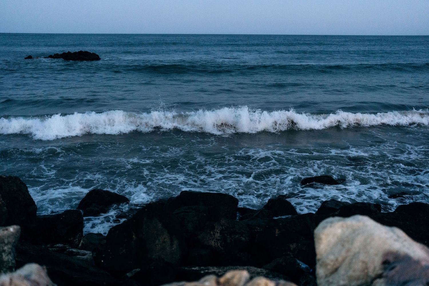 Ocean waves crash against dark rocks on a beach under blue sky.