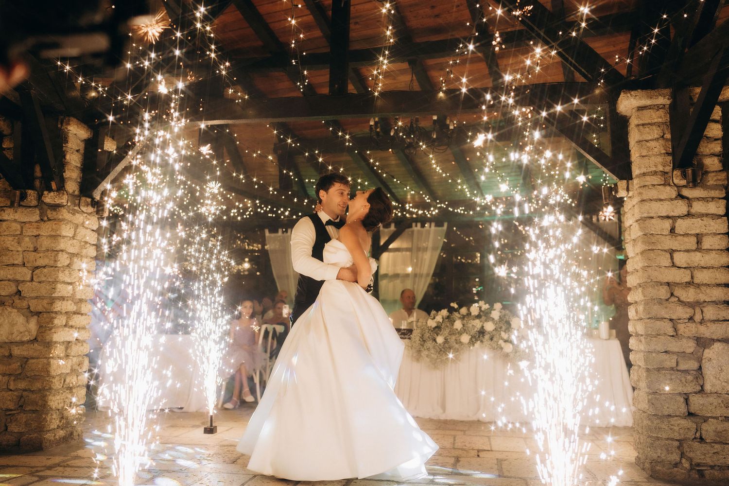 Wedding couple dancing under sparklers and string lights in a rustic stone venue.