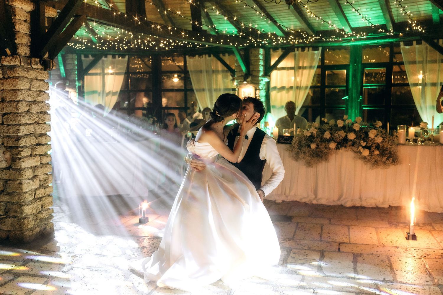 Wedding couple dancing under green-lit string lights in a rustic venue.