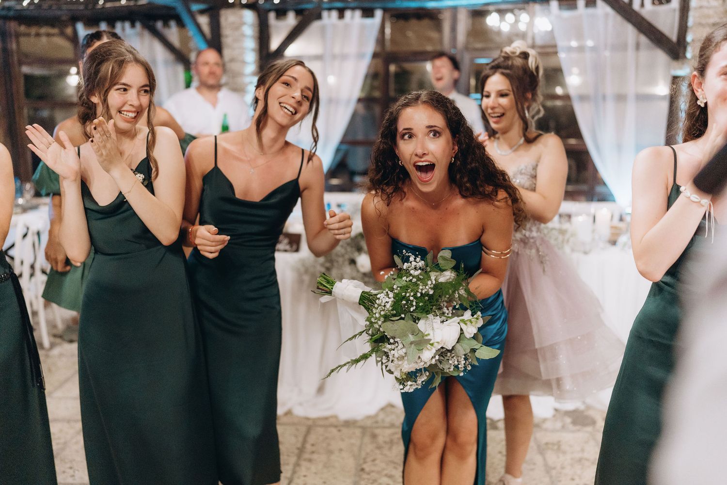 Wedding party in dark green dresses reacting with joy during reception celebrations.