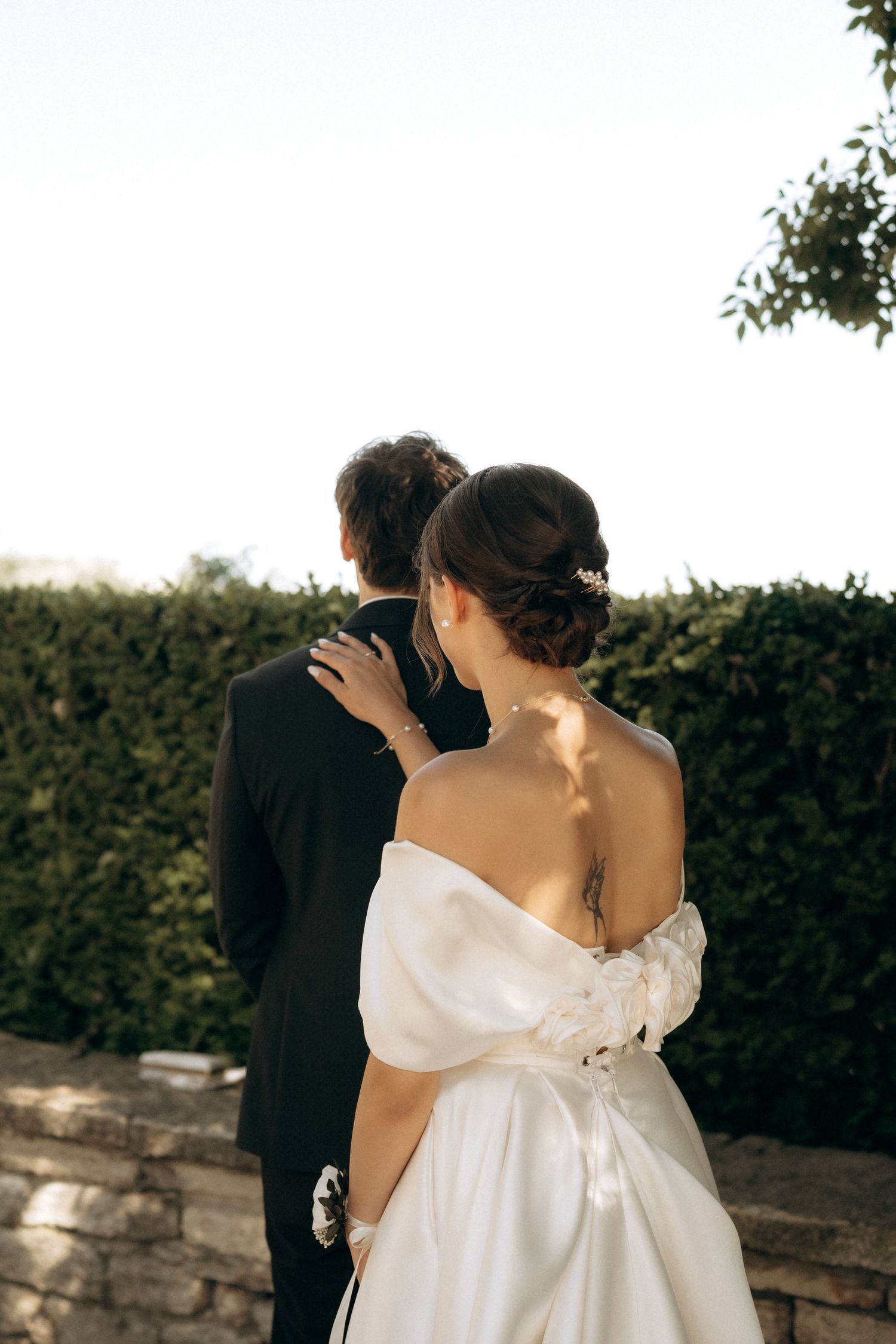 Romantic wedding couple poses against stone wall and hedges in garden setting.