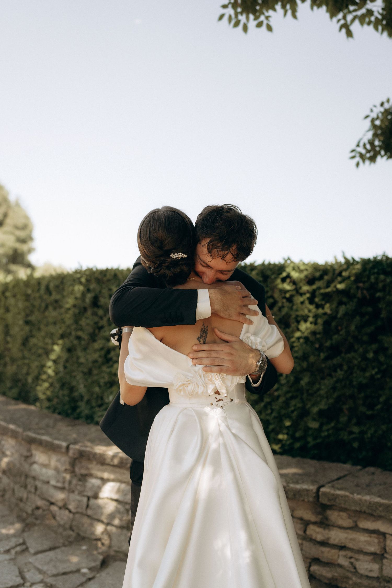 A tender embrace between newlyweds in formal attire by a garden wall.
