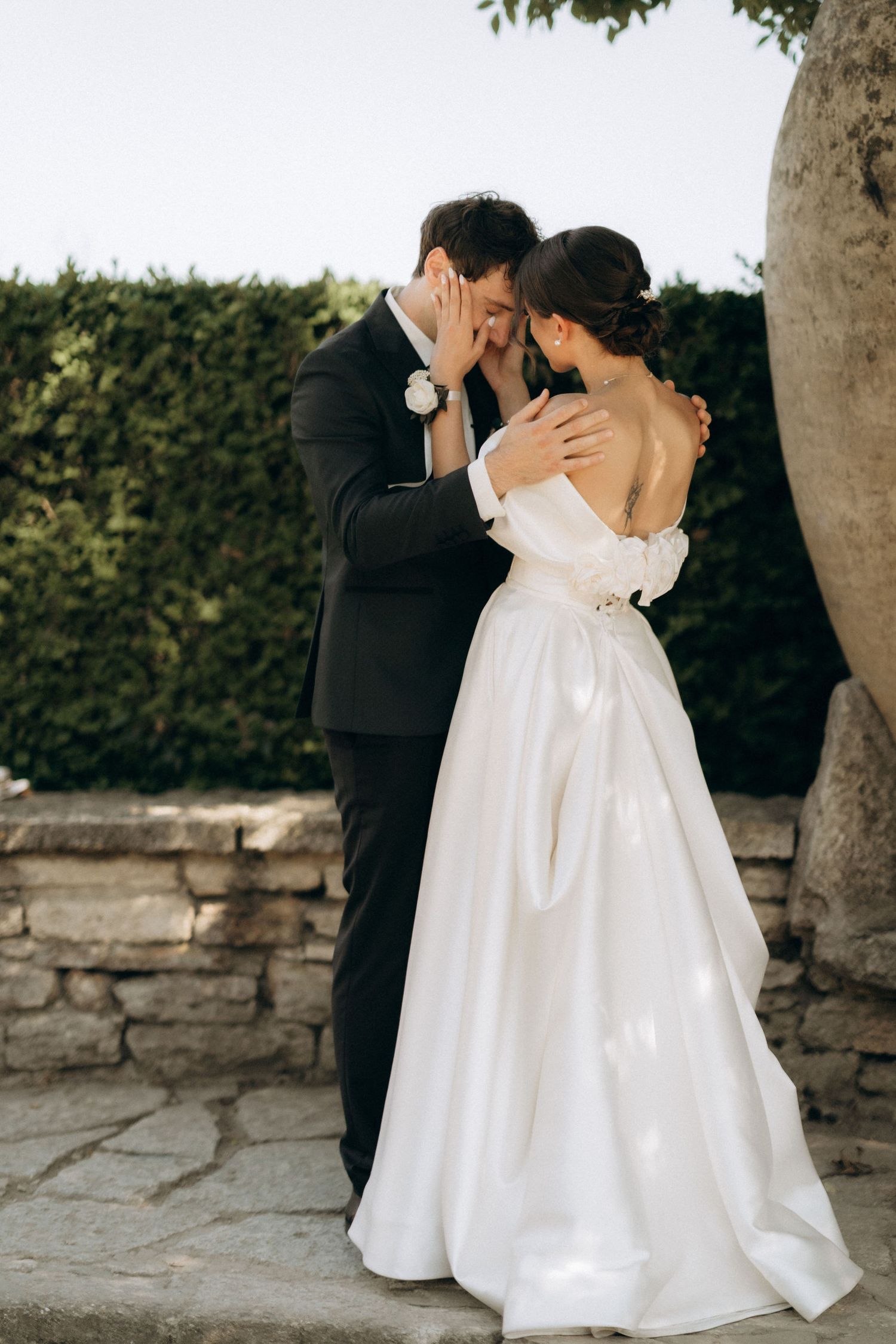 A romantic wedding scene with a couple embracing near stone walls and greenery.