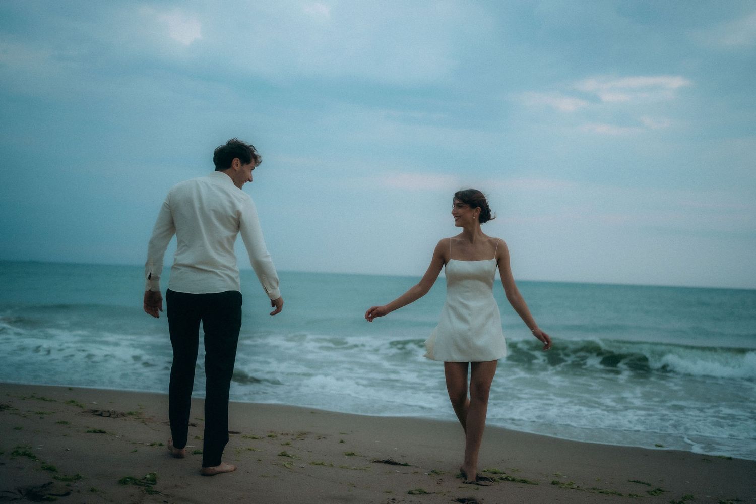 Couple playfully walking along beach with waves crashing behind them.