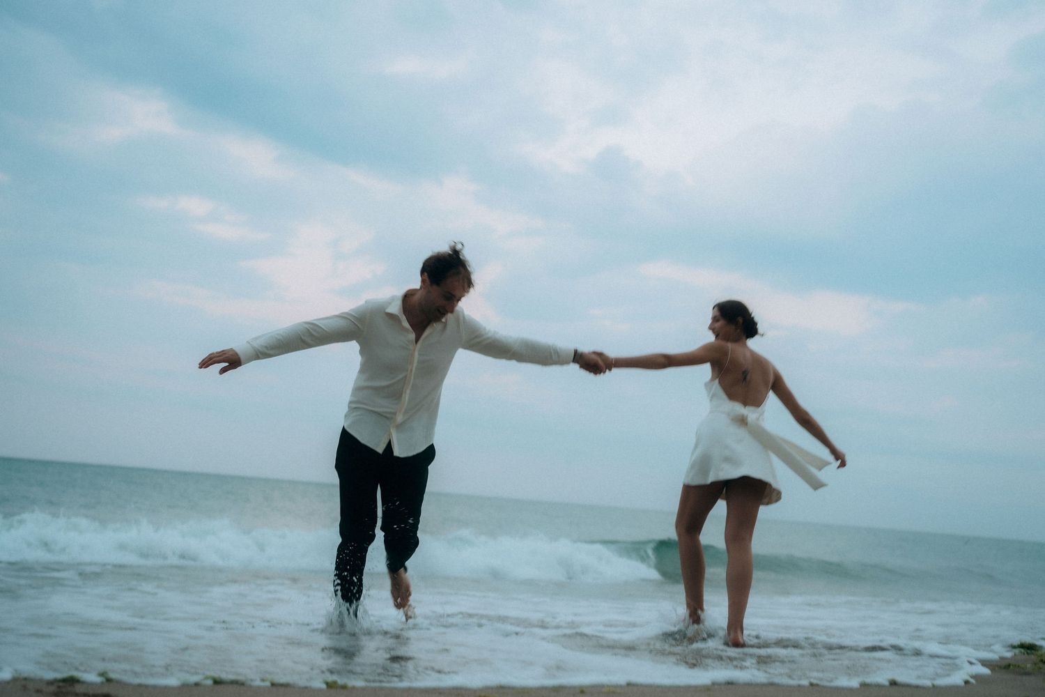 Couple playing and dancing together on a scenic beach with crashing waves and cloudy skies.