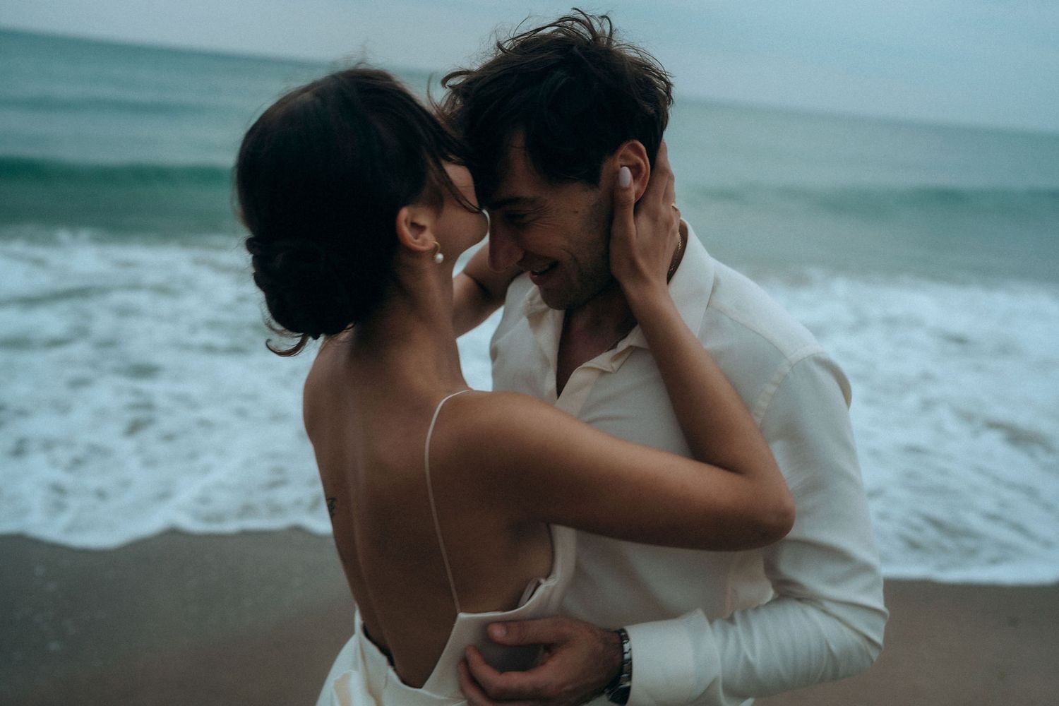 Romantic beach photoshoot featuring couple in white clothing against ocean backdrop.