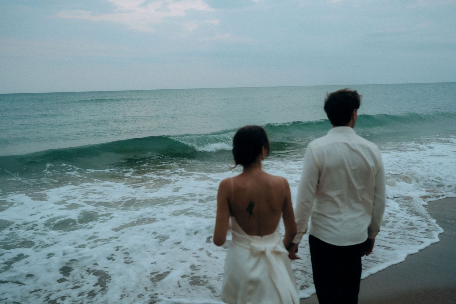 Couple walking hand in hand along shoreline with waves crashing behind them.
