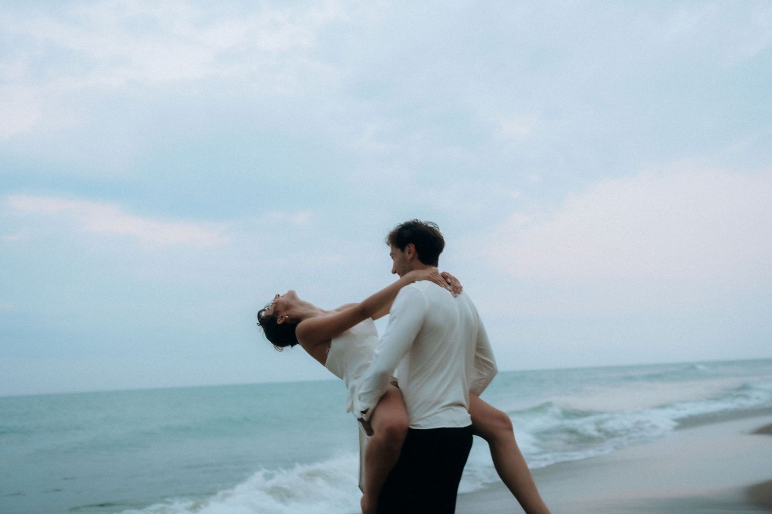 Couple dancing and dipping dramatically against cloudy beach backdrop.