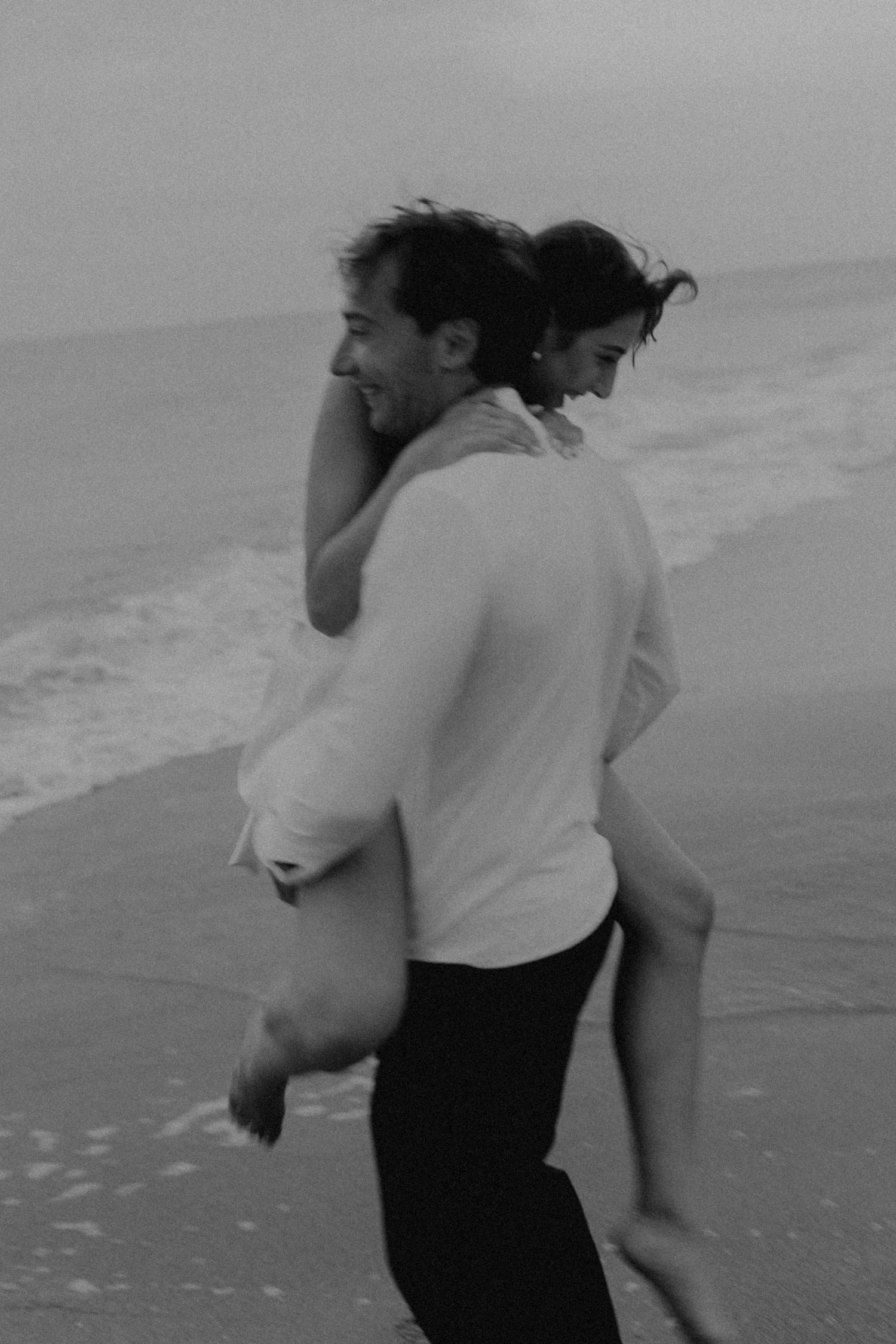 A couple sharing a playful piggyback ride moment along a misty beach in black and white.