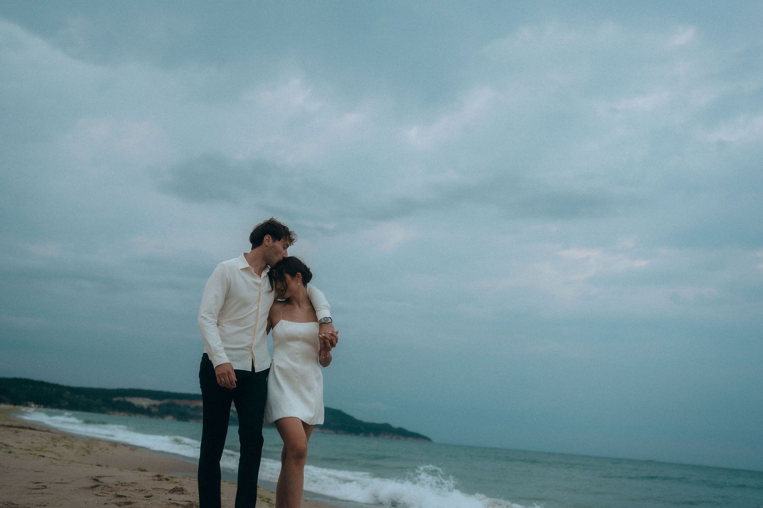 Wide shot of couple embracing on empty beach with stormy seas in background.