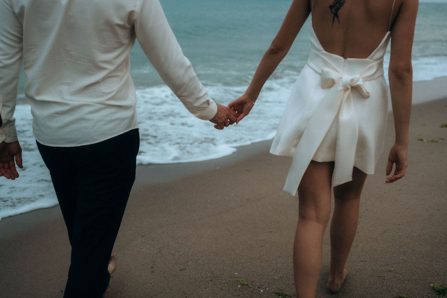 Close-up view of a couple holding hands while walking on the beach, focusing on white dress and formal attire.