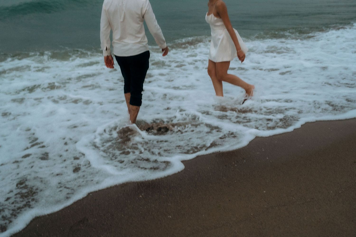 Couple wading through gentle ocean waves on a cloudy beach day.