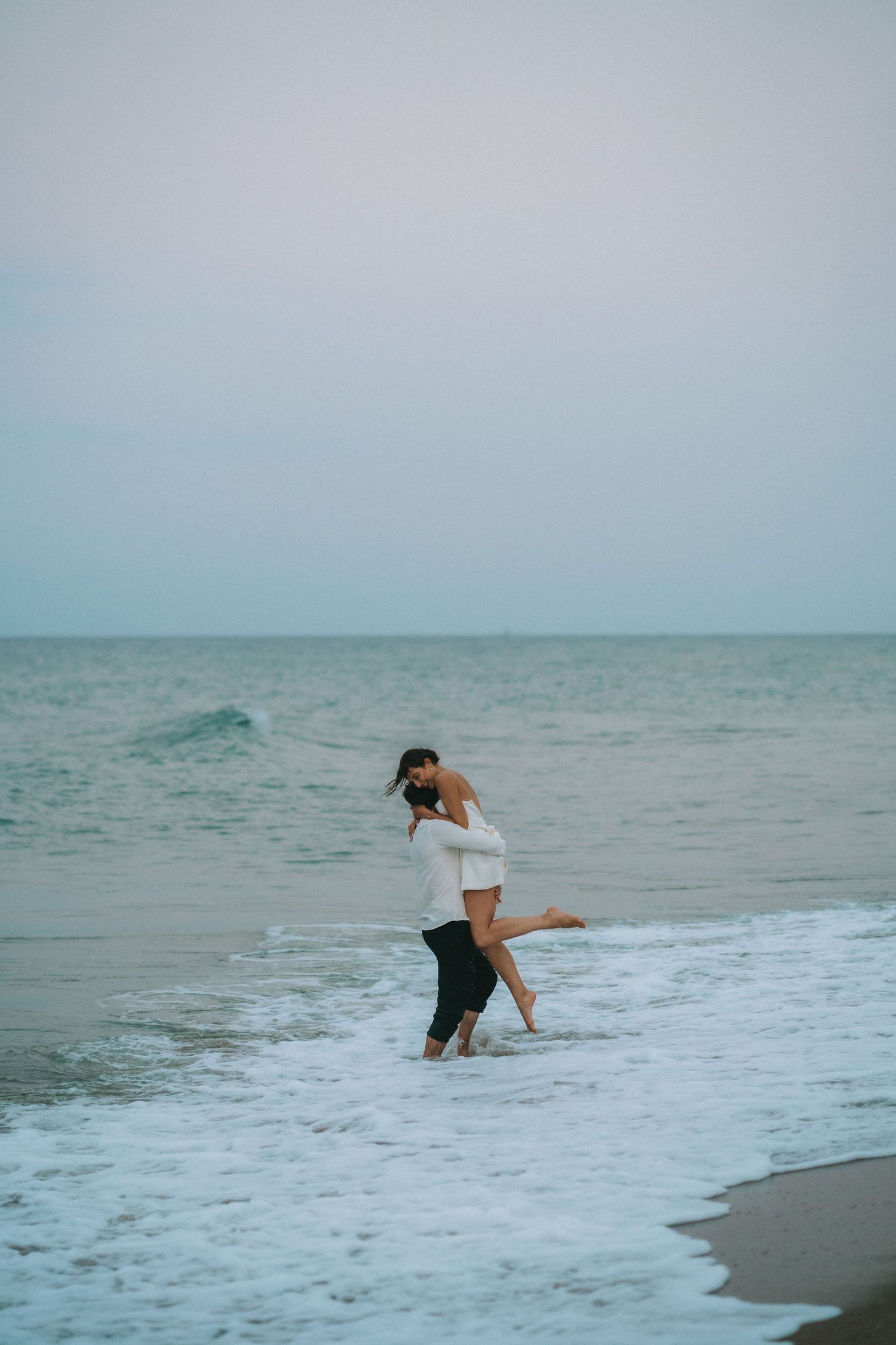 A couple plays in the ocean waves at the beach during an overcast day.