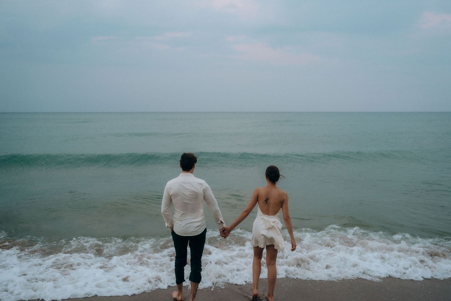 Two people walk along the shoreline as waves crash on a moody beach.