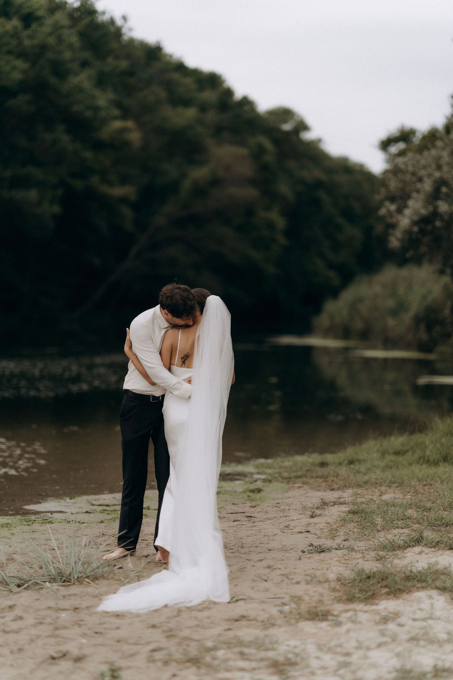 Minimalist wedding photography on sandy beach with lake and trees in background.