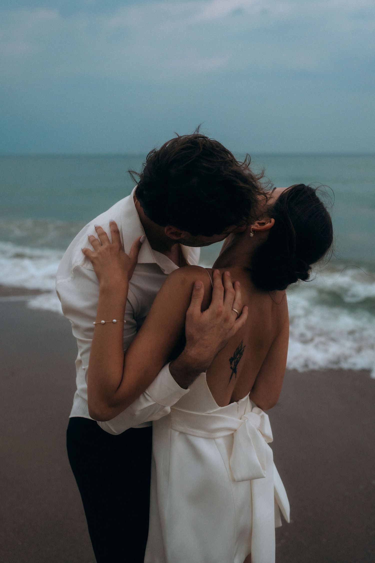 A romantic beach photoshoot featuring a couple in formal attire embracing near the water.