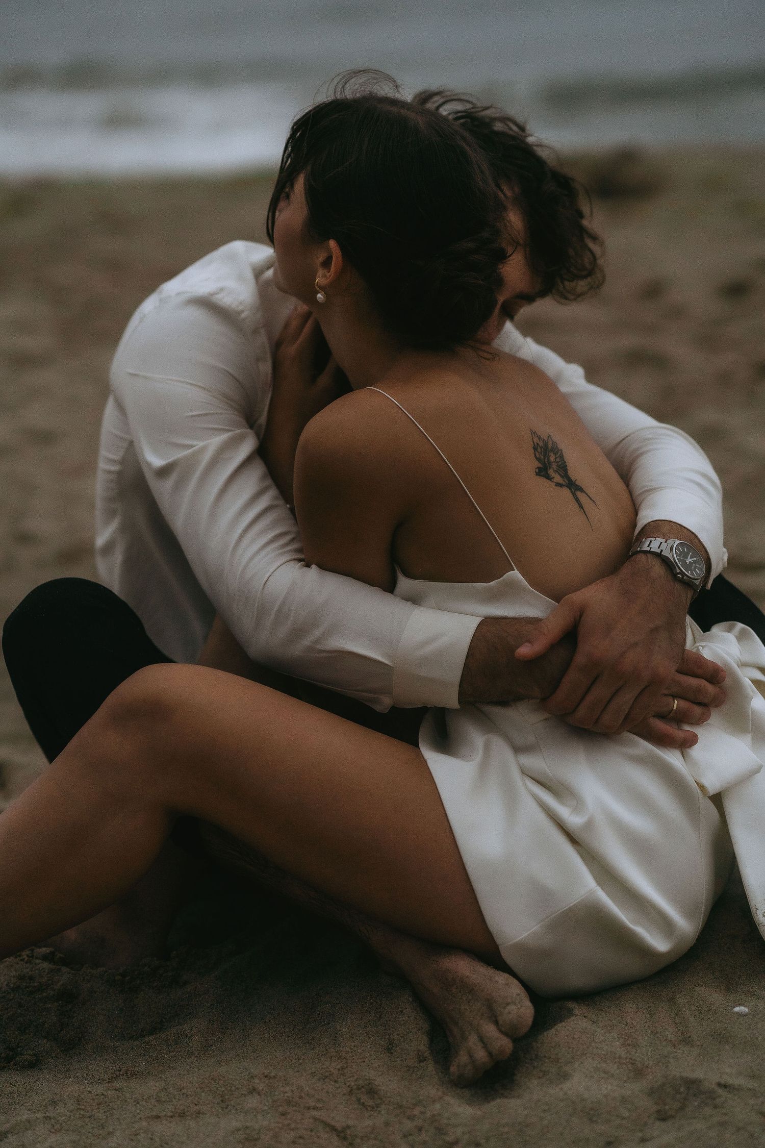 A couple in wedding attire shares tender moments while lying on the beach sand.