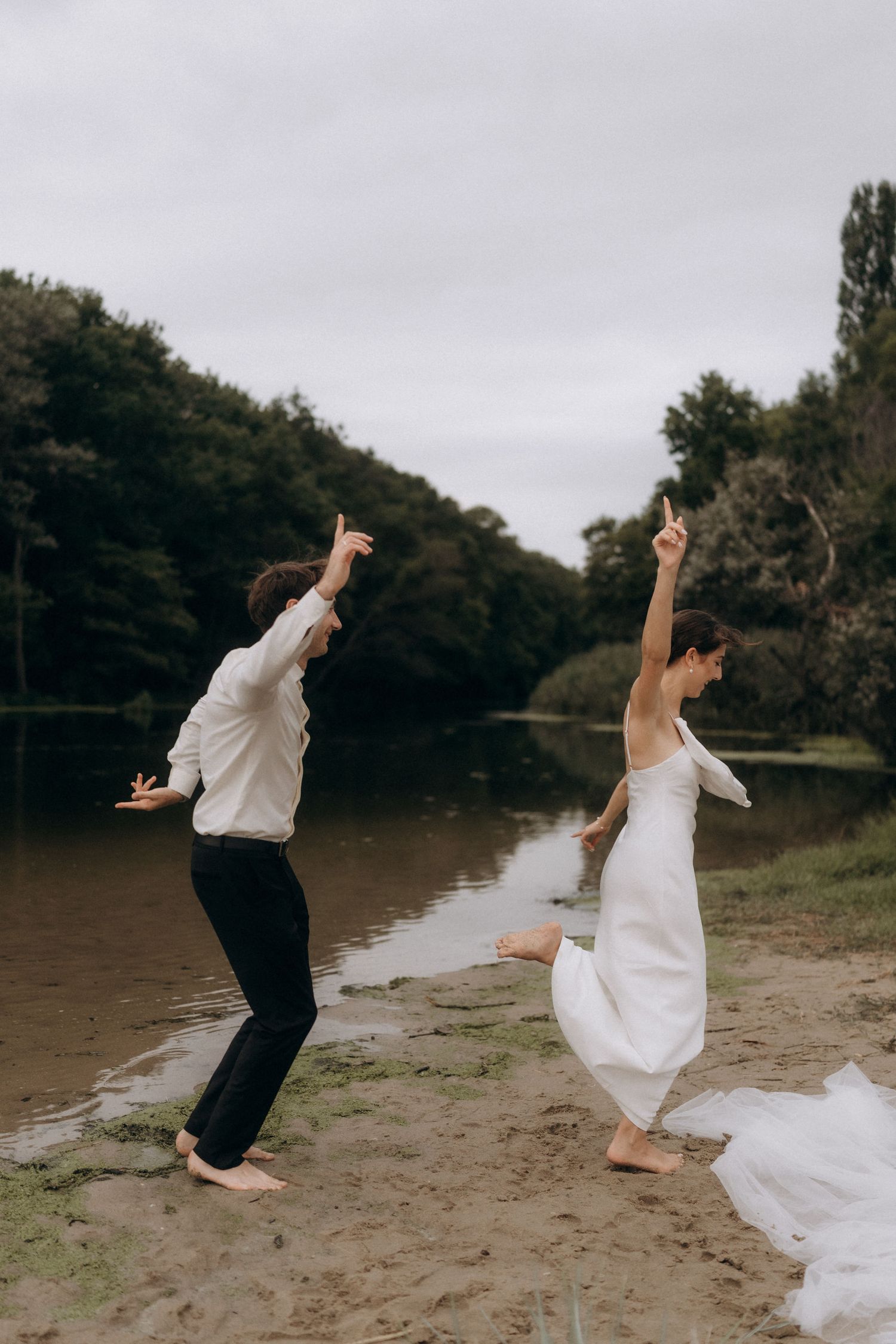 Playful wedding couple jumping and dancing on sandy beach by the lake.