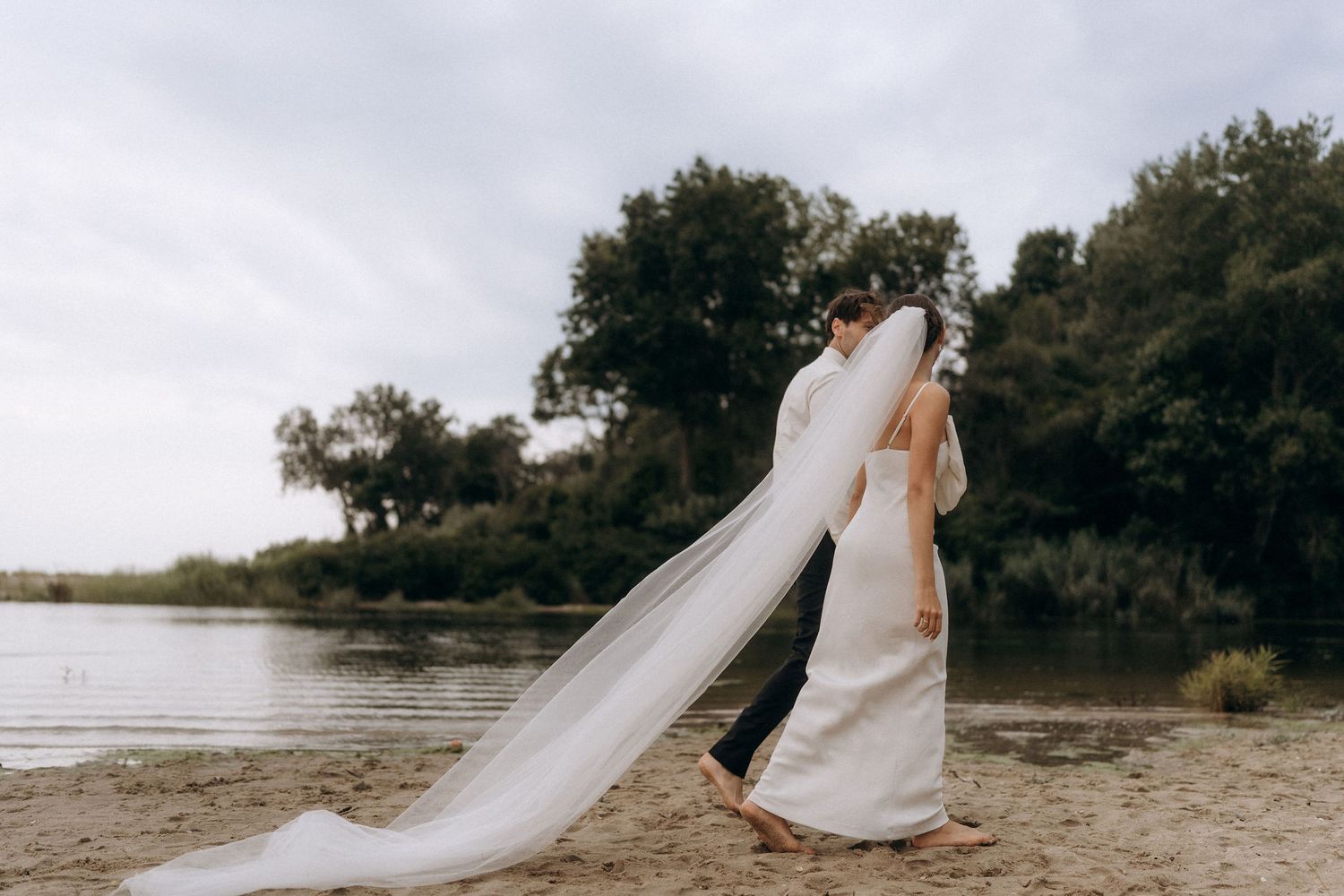 Wedding couple walking along serene lakeside with flowing veil in gentle breeze.