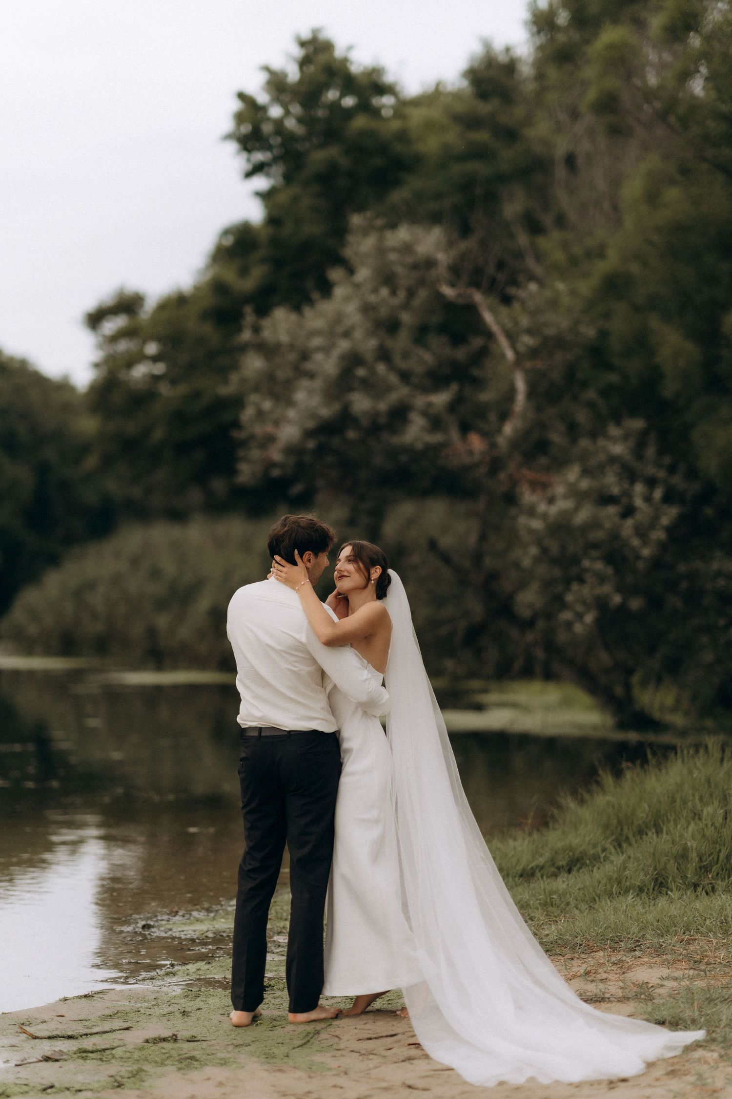 Wedding couple in white embracing by a serene lake surrounded by trees.