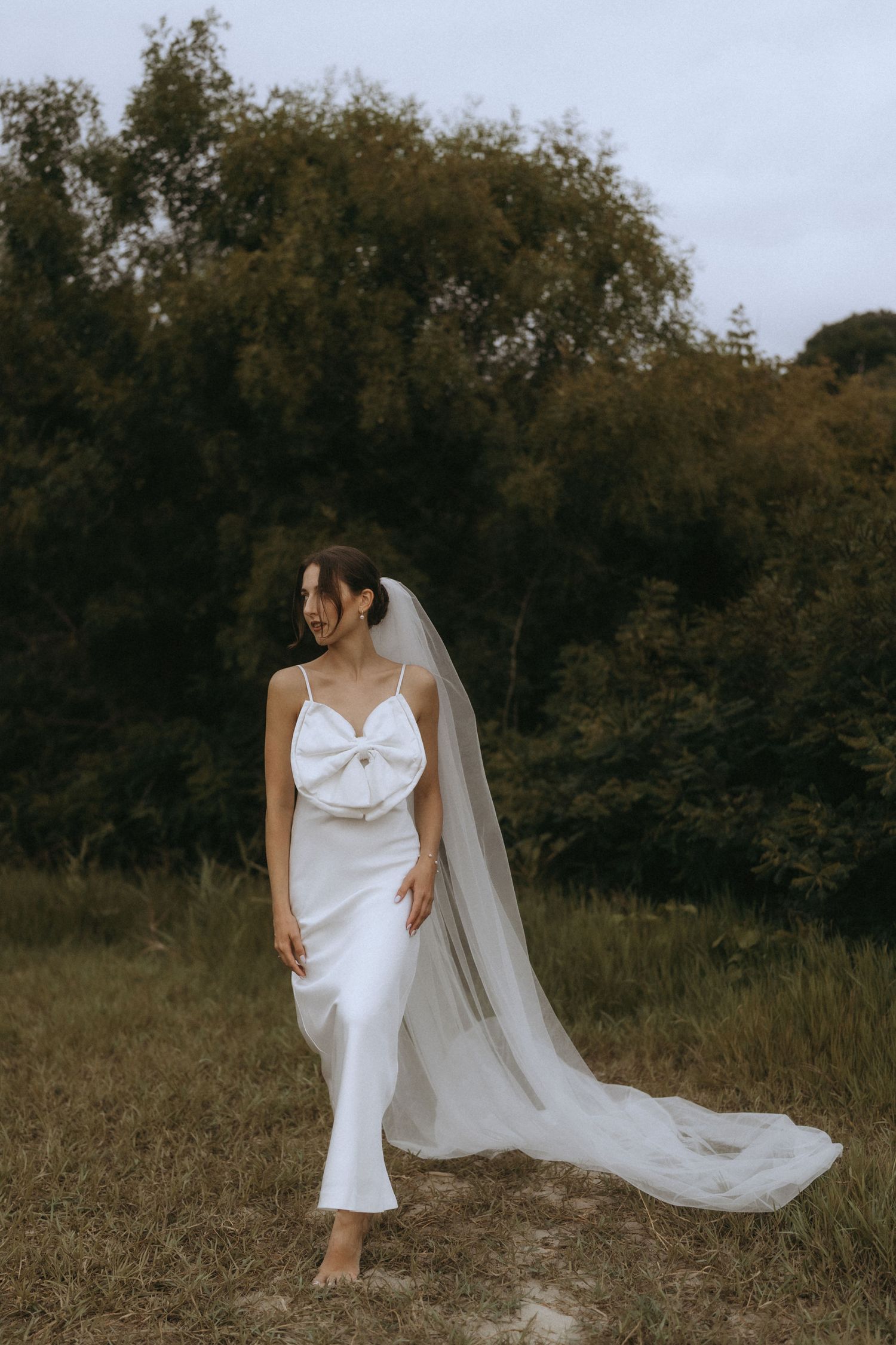 Bride in flowing white dress and veil poses gracefully in a rustic outdoor setting.