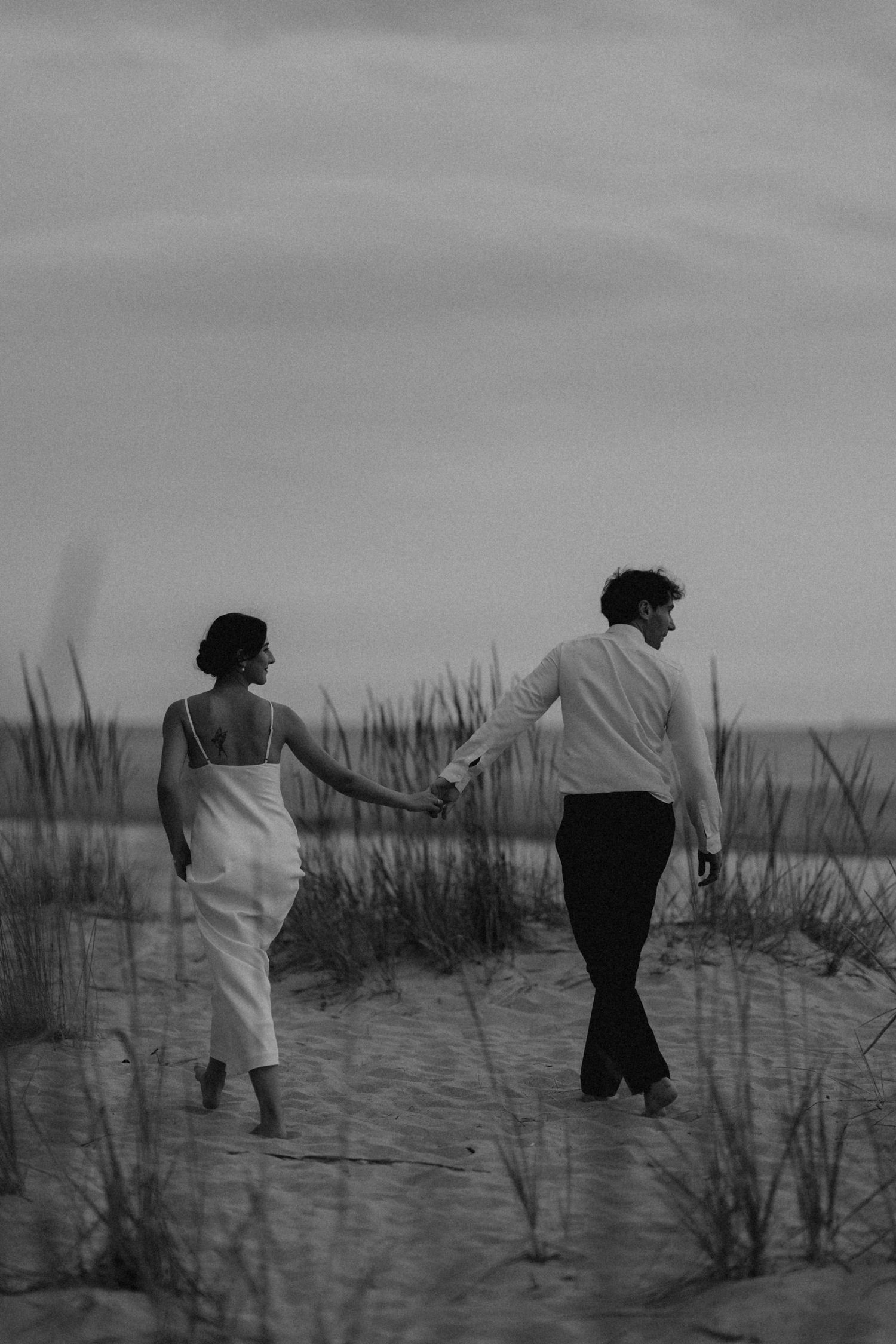 Black and white photo of couple dancing playfully among beach grass at sunset.