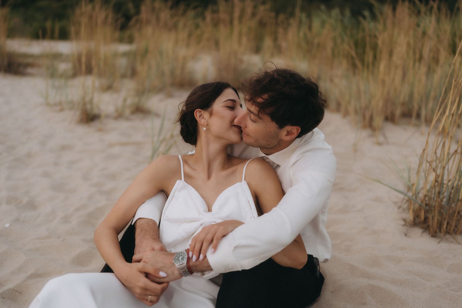 Couple embraces tenderly on beach with golden grass in background.