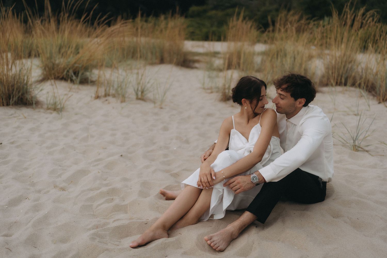 Romantic couple shares intimate moments on sandy beach with natural grass backdrop.