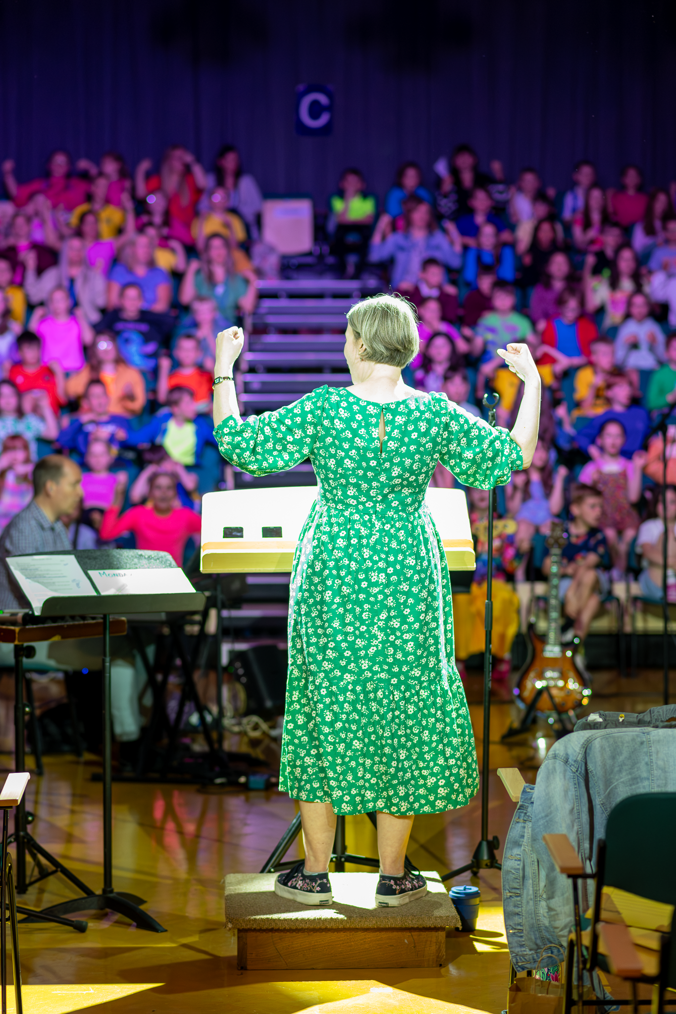 A choir conductor in a green polka dot dress leads a performance of a colourfully dressed children's choir.