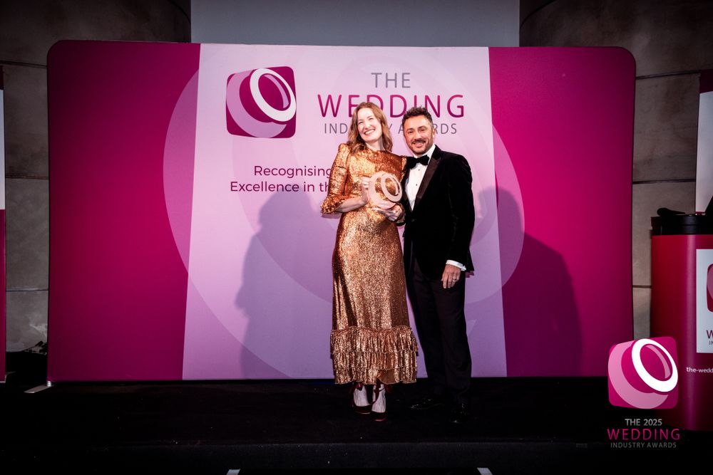 Award winner in gold dress poses with presenter on stage at The Wedding Industry Awards 2025.