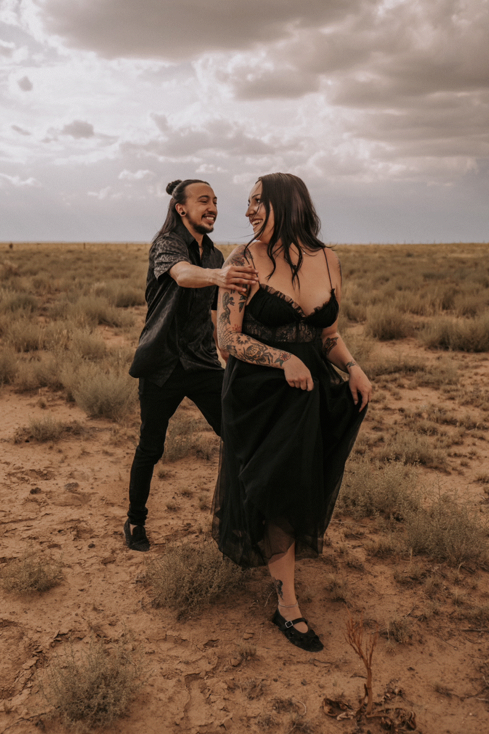 A couple dances together in a desert landscape under moody clouds at sunset. Queer engagement photographer Albuquerque. New Mexico weddings.