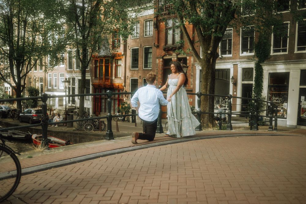 Man proposes on one knee to woman in white dress on cobblestone street in Amsterdam.