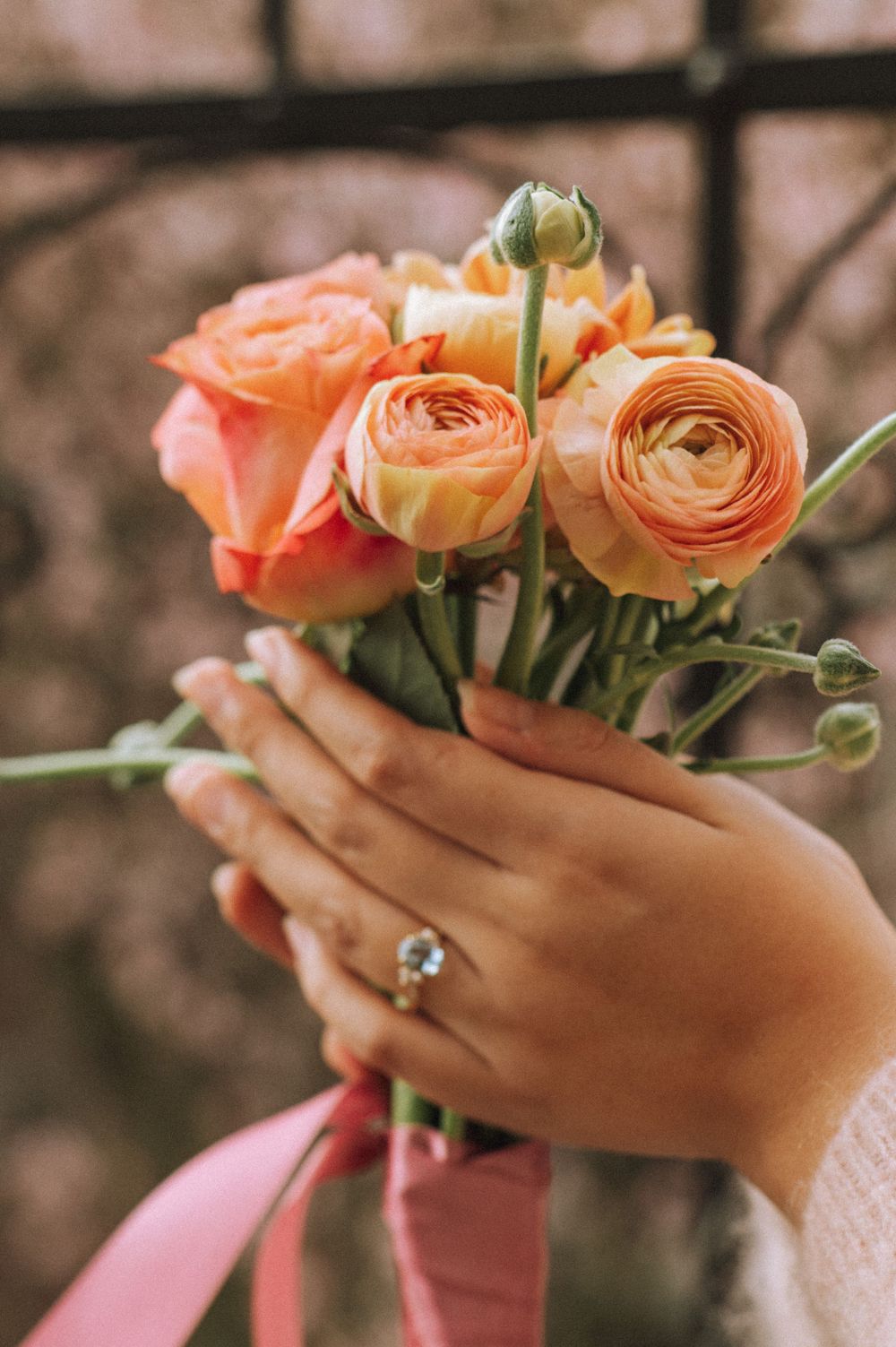 Hands holding delicate peach-colored roses with an engagement ring visible.